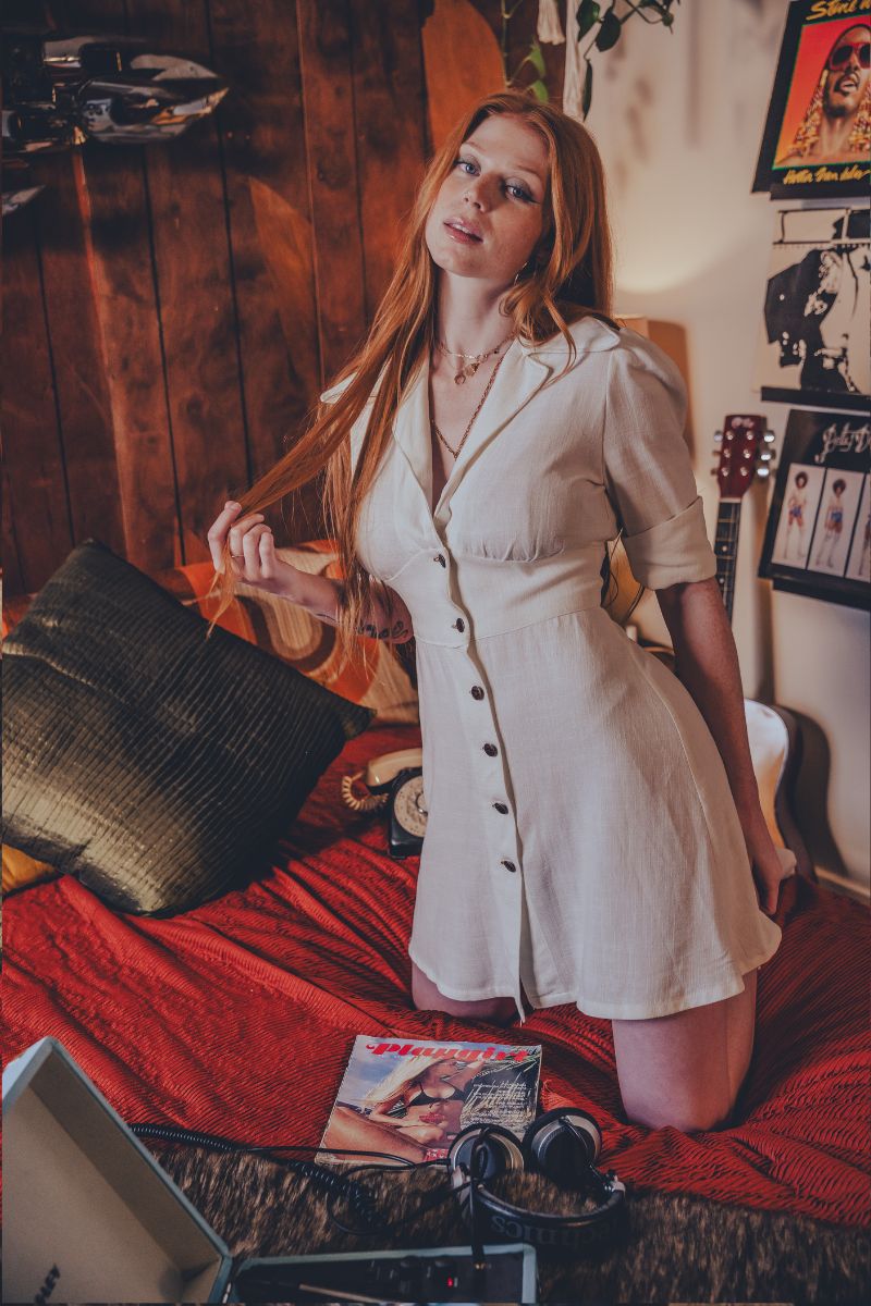 Woman in a white dress sitting on a bed in a room with wooden walls and posters.
