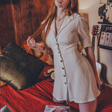 Woman in a white dress sitting on a bed in a room with wooden walls and posters.