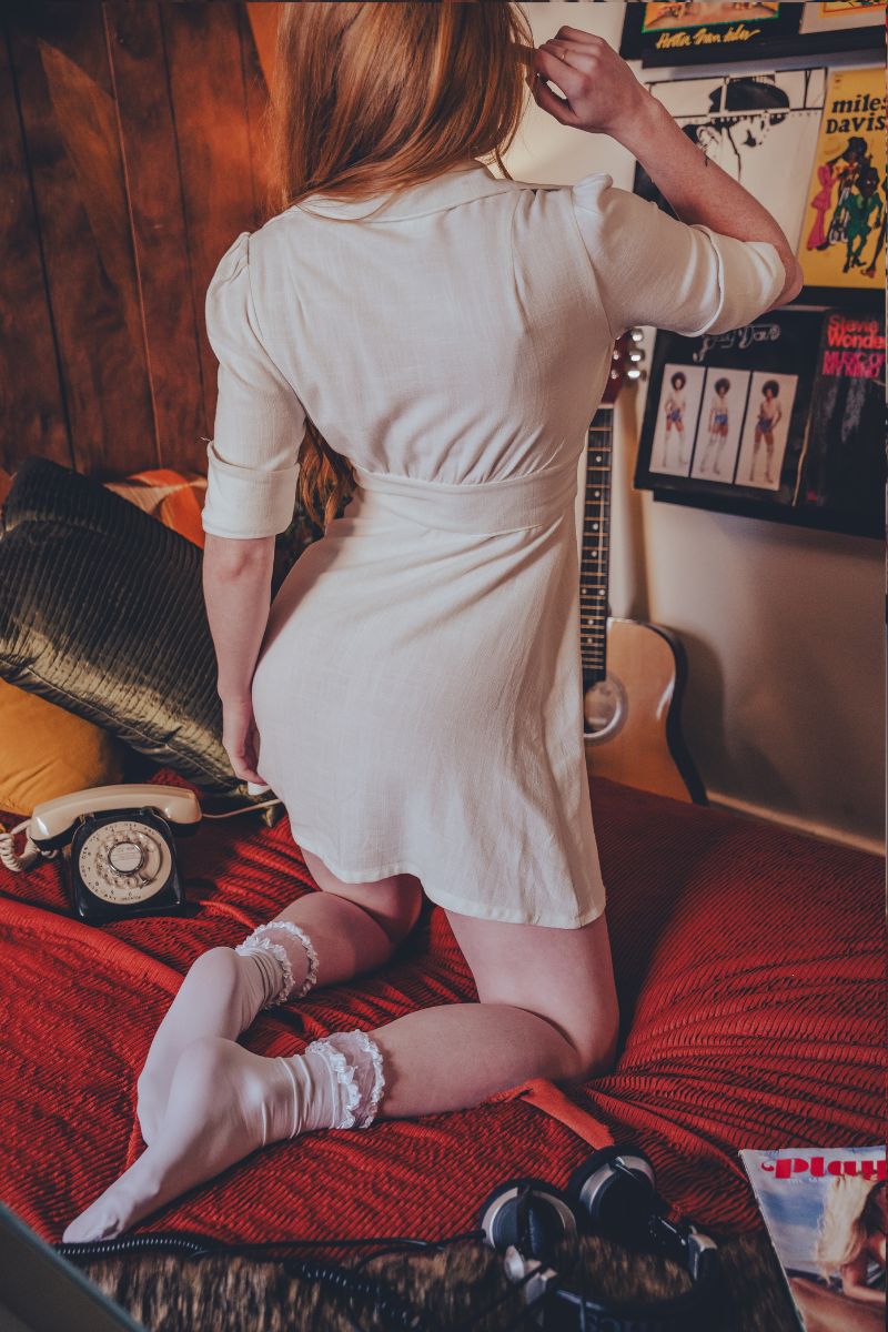 Person in a white dress standing on a bed with a guitar and posters in the background