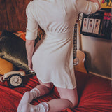 Person in a white dress standing on a bed with a guitar and posters in the background