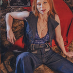 Woman sitting on a patterned rug wearing a sleeveless denim vest and jeans, with a red couch and guitar in the background.