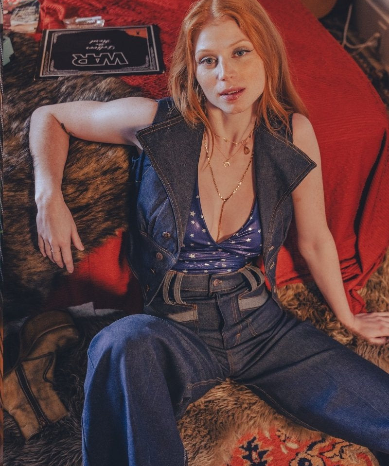 Woman sitting on a patterned rug wearing a sleeveless denim vest and jeans, with a red couch and guitar in the background.
