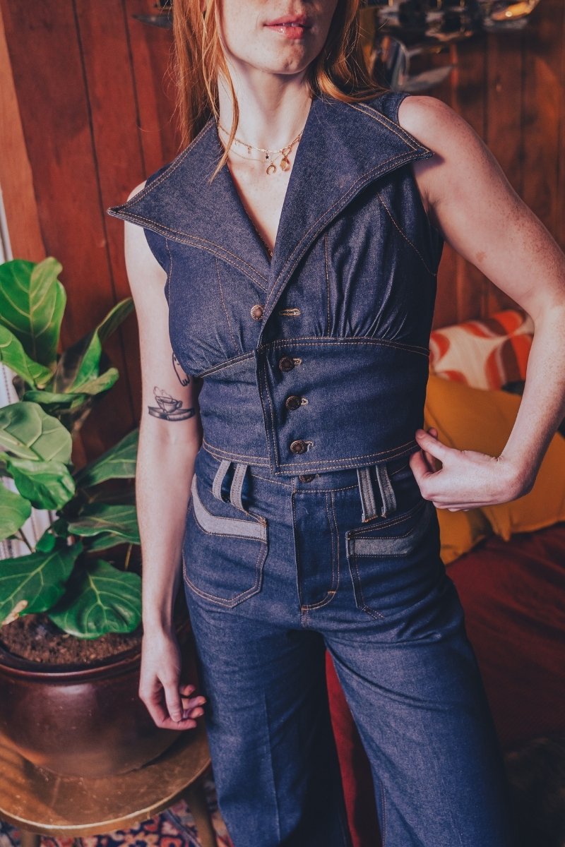 Person wearing a 1970s inspired denim jumpsuit standing indoors with a plant and groovy furniture in the background