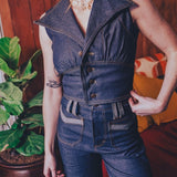 Person wearing a 1970s inspired denim jumpsuit standing indoors with a plant and groovy furniture in the background