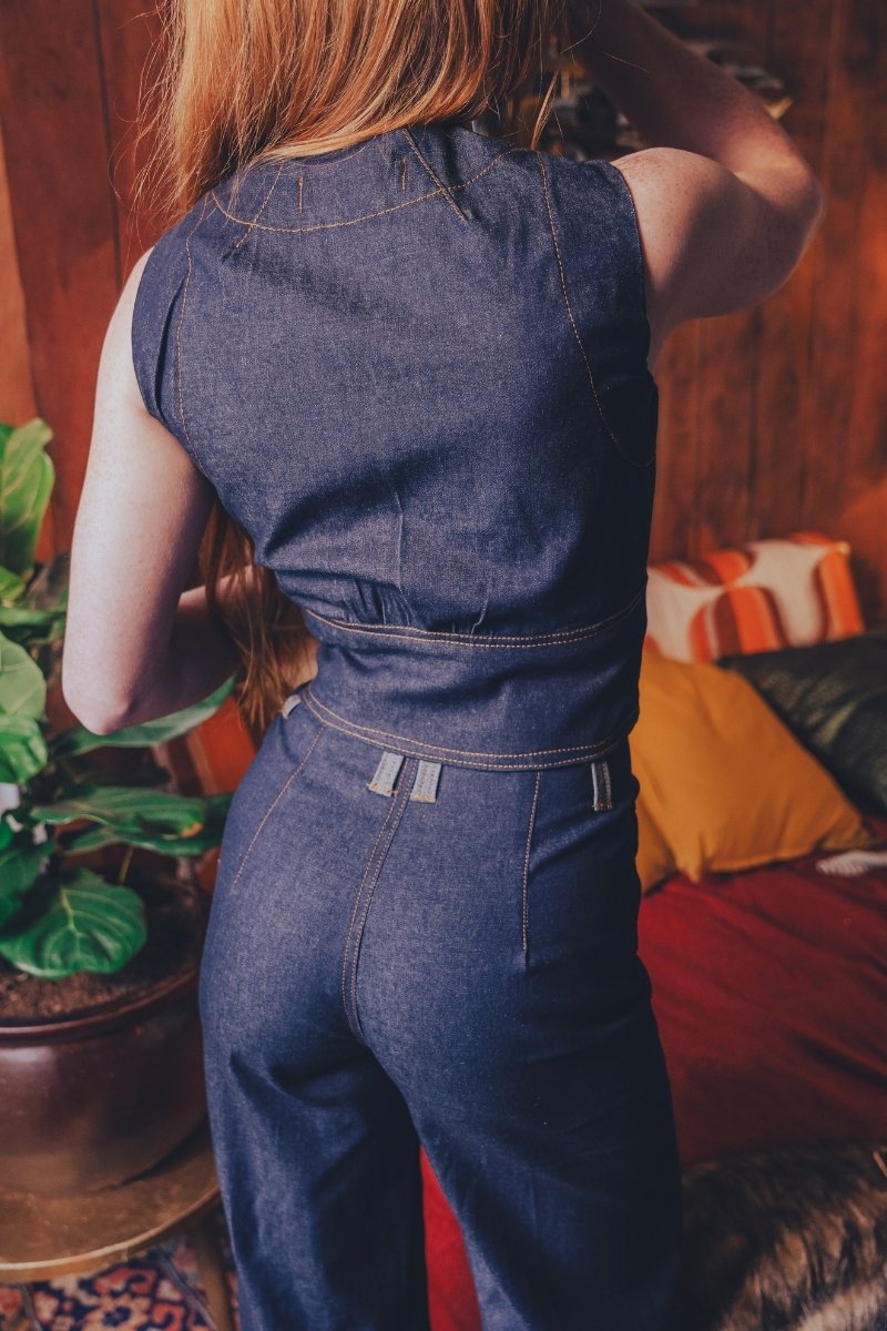 Person wearing a blue denim 70s inspired jumpsuit in an funky indoor setting with a plant and colorful cushions.