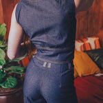 Person wearing a blue denim 70s inspired jumpsuit in an funky indoor setting with a plant and colorful cushions.