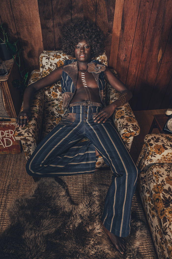 A person sitting in a room with a wooden background, wearing a blue and white striped waistcoat with a brooch, paired with hipster bells. The room also features a furry rug and a bookshelf with books.