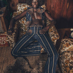 A person sitting in a room with a wooden background, wearing a blue and white striped waistcoat with a brooch, paired with hipster bells. The room also features a furry rug and a bookshelf with books.