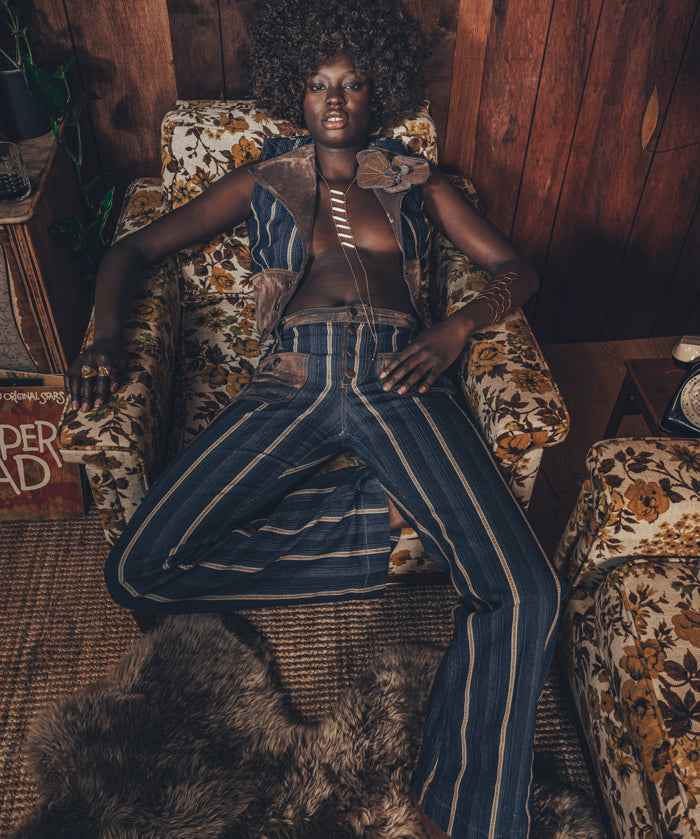 A person sitting in a room with a wooden background, wearing a blue and white striped waistcoat with a brooch, paired with hipster bells. The room also features a furry rug and a bookshelf with books.