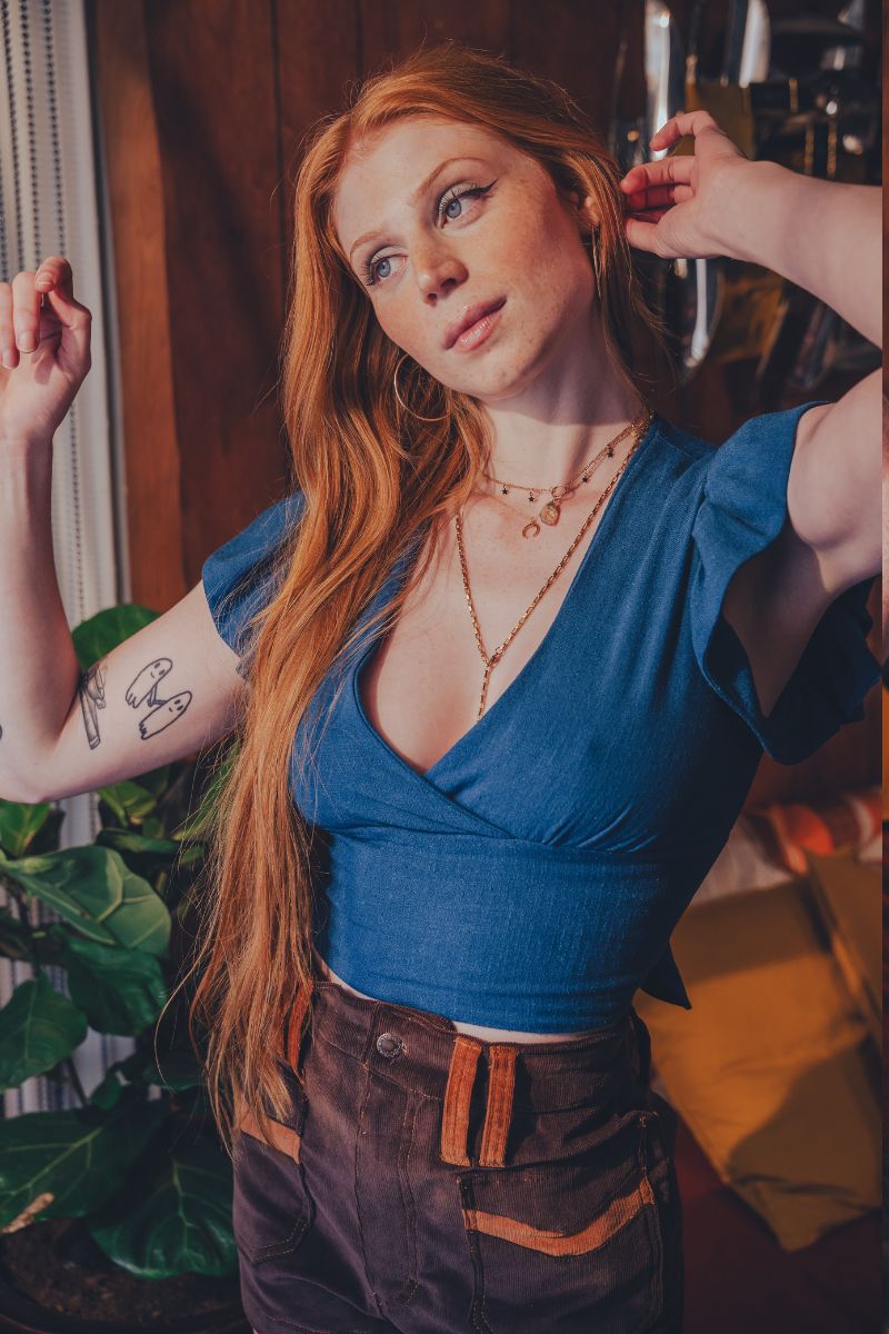 Woman in a blue top and brown pants posing indoors with plants and furniture in the background