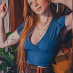 Woman in a blue top and brown pants posing indoors with plants and furniture in the background