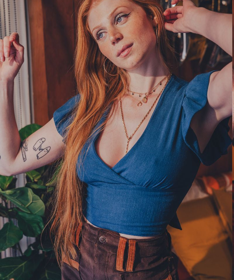 Woman in a blue top and brown pants posing indoors with plants and furniture in the background