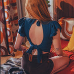 Woman sitting on a bed wearing a blue top with an open back, in a room with patterned curtains and a red blanket.