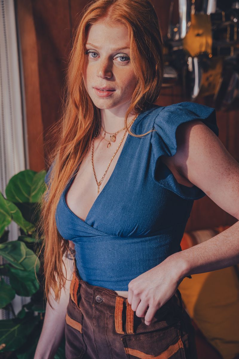 Woman with long red hair wearing a blue top and brown pants in an indoor setting.