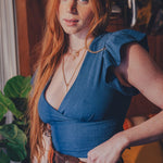 Woman with long red hair wearing a blue top and brown pants in an indoor setting.