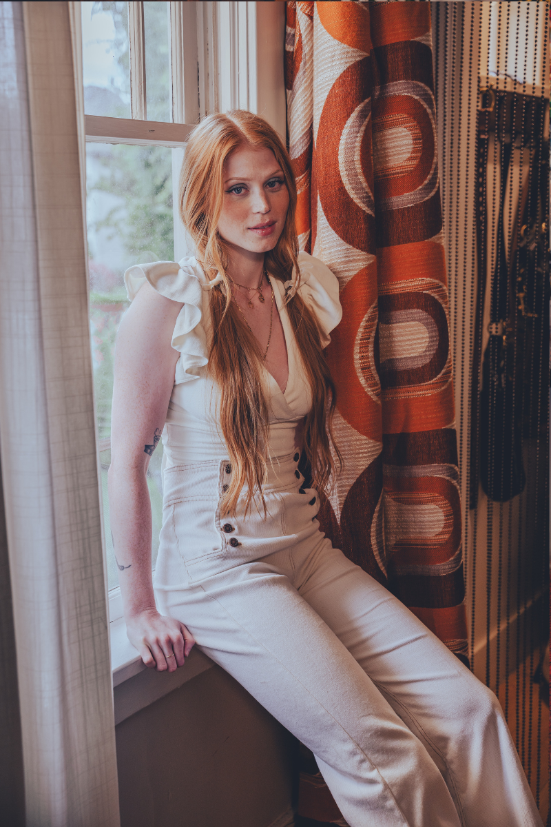 Woman in a white sexy 70s bell bottom outfit sitting by a window with a retro 1970s patterned curtain.