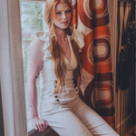 Woman in a white sexy 70s bell bottom outfit sitting by a window with a retro 1970s patterned curtain.