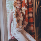 Woman in a white sexy 70s bell bottom outfit sitting by a window with a retro 1970s patterned curtain.