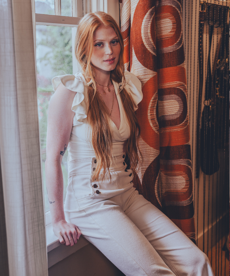 Woman in a white sexy 70s bell bottom outfit sitting by a window with a retro 1970s patterned curtain.
