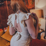 Person wearing a white  boho backless top with ruffled sleeves in a room with 1970s style wooden walls and a guitar.