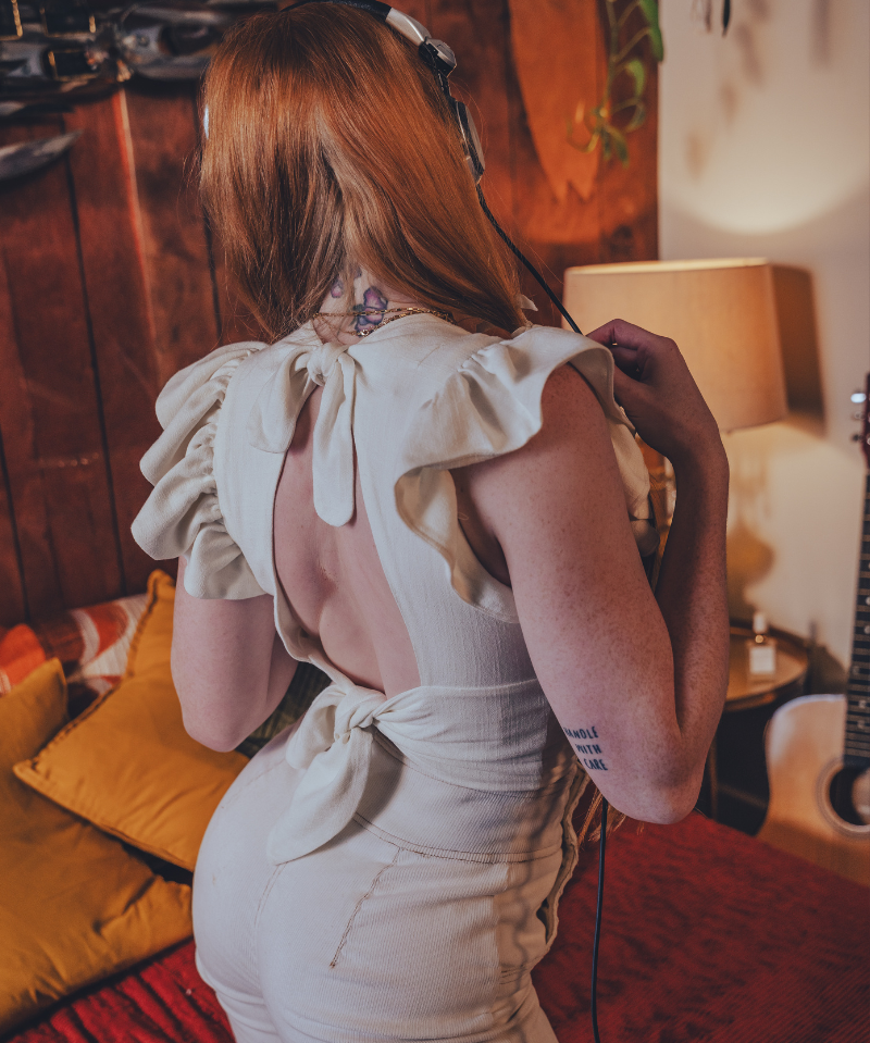 Person wearing a white  boho backless top with ruffled sleeves in a room with 1970s style wooden walls and a guitar.