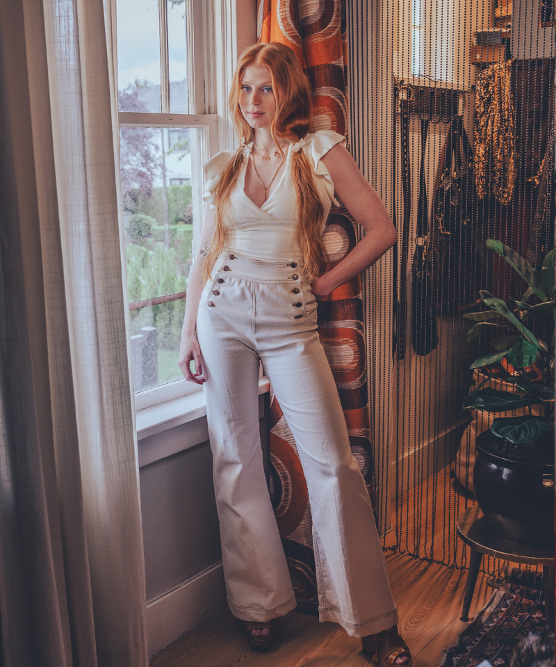 Woman in white bellbottom jeans in a room with a window and vintage style 1970s decorative items.