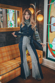 A person wearing a retro denim jumpsuit with long sleeves, seated on a funky surface, with a guitar and wooden decor in the background.