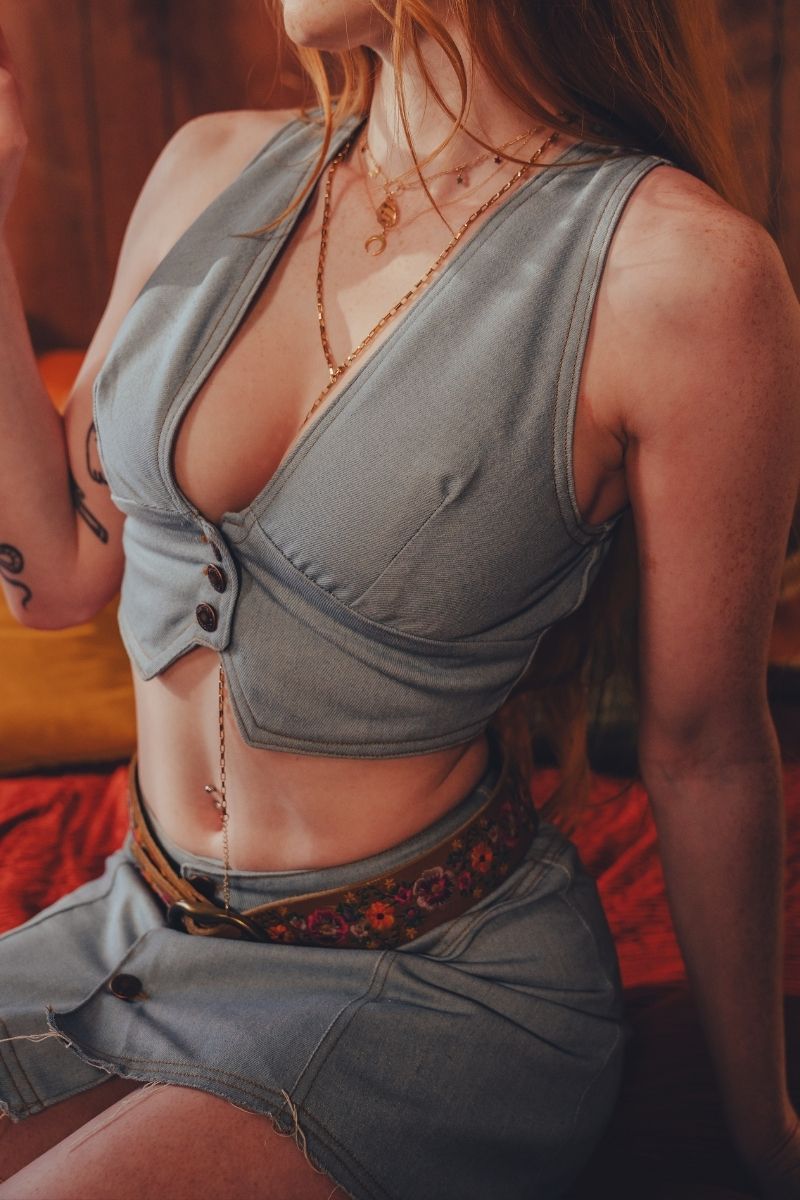 Person wearing a faded denim 70s inspired waistcoat  with a woodstock inspired floral belt in a warm-toned  1970s setting
