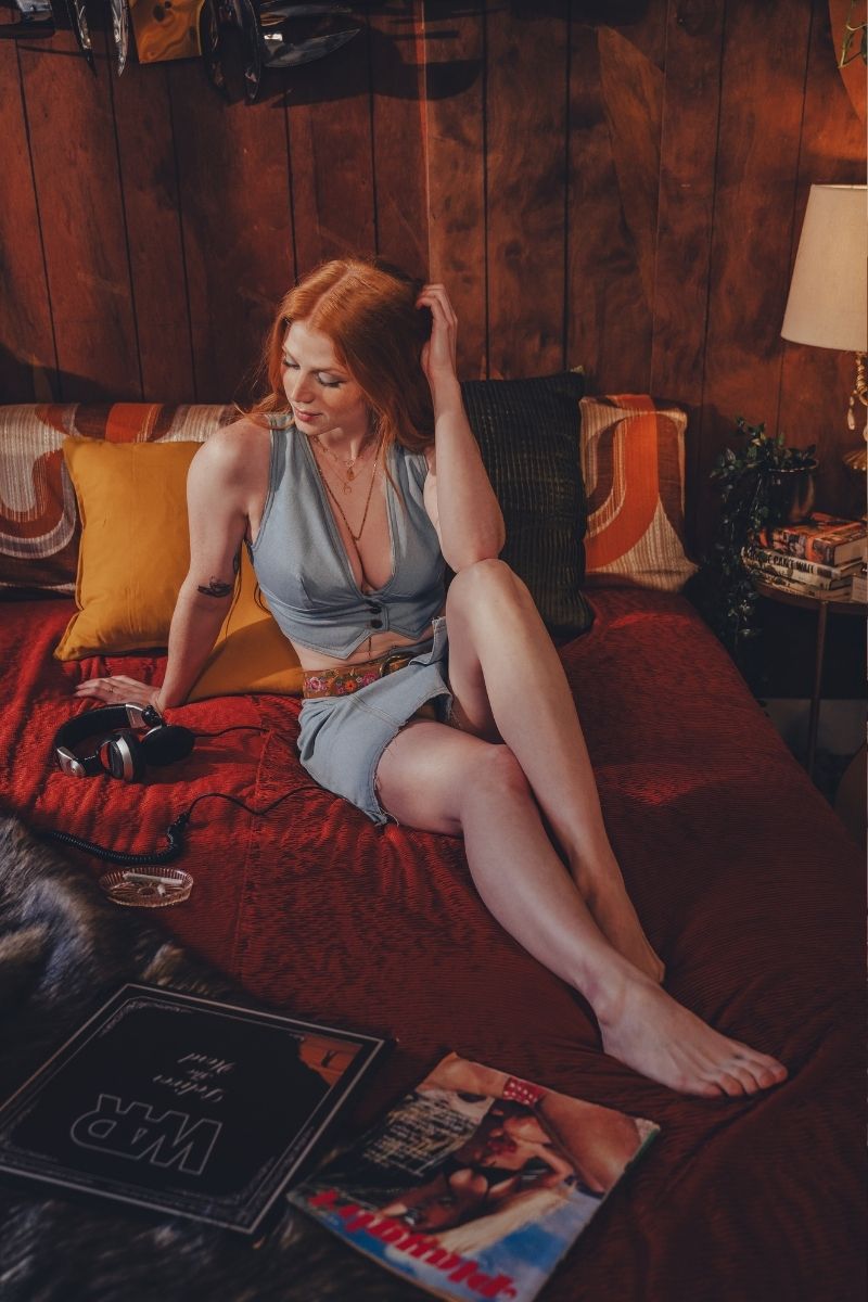 Woman sitting on a 70's style bed in a cozy room with a lamp and book wearing wood stock inspired festival mini skirt and waistcoat in denim