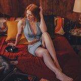 Woman sitting on a 70's style bed in a cozy room with a lamp and book wearing wood stock inspired festival mini skirt and waistcoat in denim