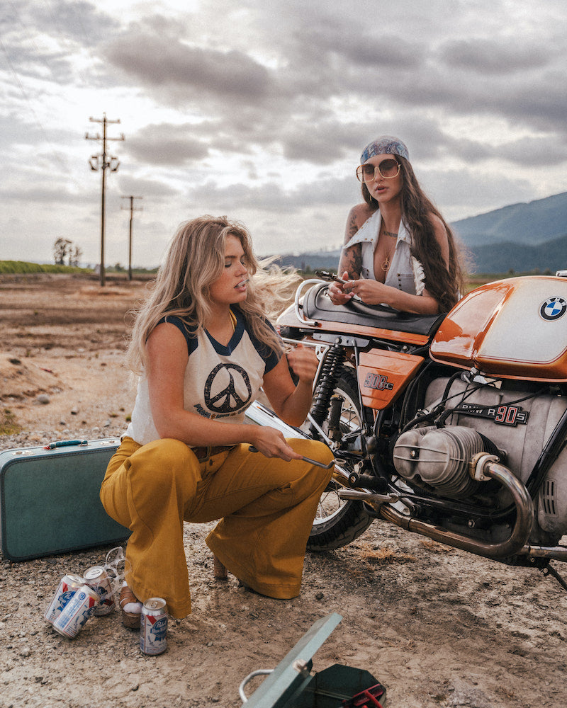 Two women sitting next to a motorcycle in an open area with mountains in the background wearing Soul Sister since 1969.
