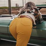 Person in yellow pants with a cow on their back, standing next to a green car in an open field wearing soul sister since 1969.