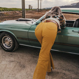 Person wearing mustard yellow flared pants standing next to a green car in an open field wearing soul sister since 1969.