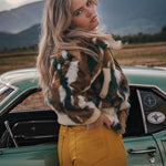 Woman in a patterned jacket and yellow pants standing next to a vintage car with mountains in the background wearing Soul Sister since 1969
