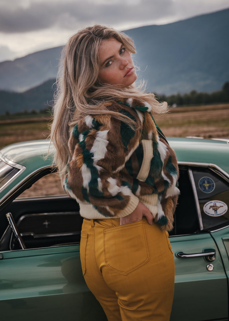 Woman in a patterned jacket and yellow pants standing next to a vintage car with mountains in the background v