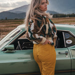 Woman in a patterned jacket and yellow pants standing next to a vintage car with mountains in the background wearing Soul Sister since 1969