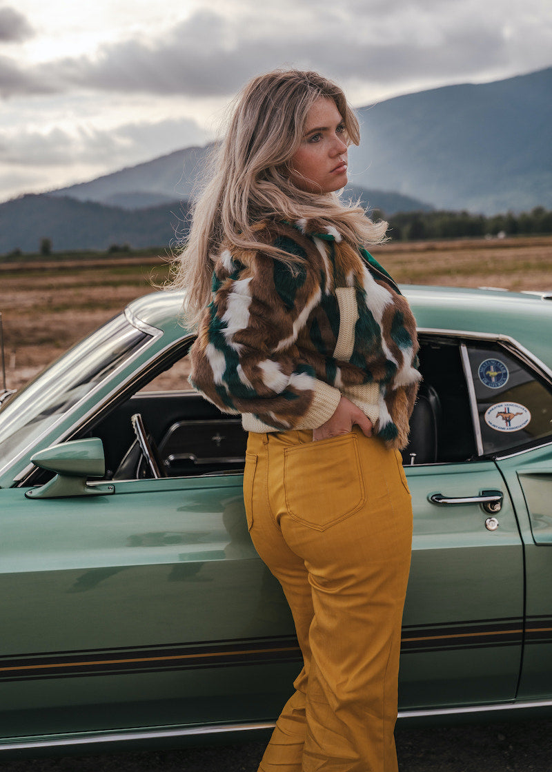 Woman in a patterned jacket and yellow pants standing next to a vintage car with mountains in the background v
