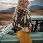 Woman in a patterned coat and yellow pants standing next to a vintage car with mountains in the background wearing Soul Sister since 1969