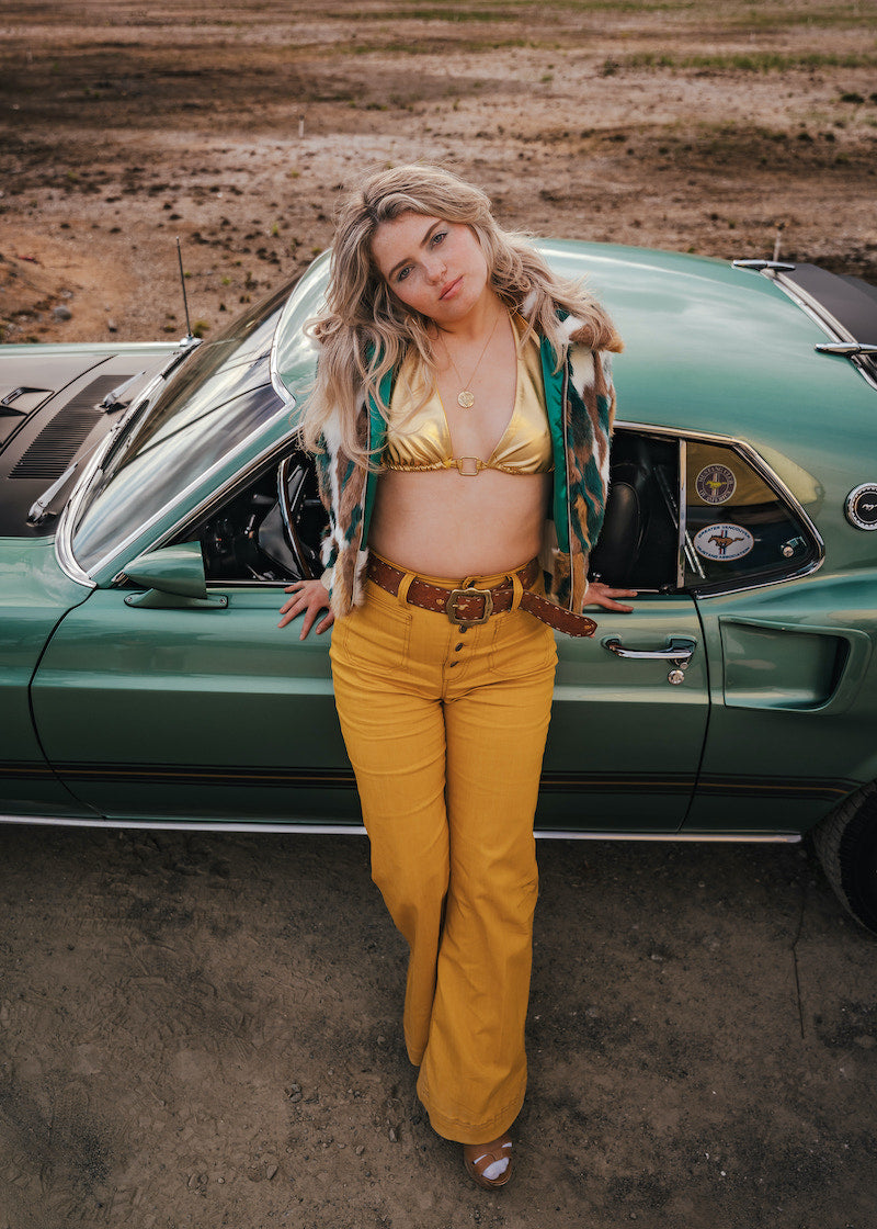 Woman in a gold bikini top and yellow pants standing next to a vintage car in a desert setting wearing soul sister since 1969