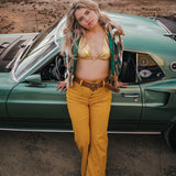 Woman in a gold bikini top and yellow pants standing next to a vintage car in a desert setting wearing soul sister since 1969