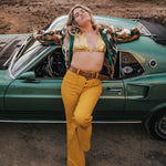 Woman in a gold bikini top and yellow pants leaning against a green vintage car in a desert setting wearing Soul Sister since 1969.