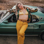 Woman in a gold top and yellow pants standing next to a vintage car in an open field wearing soul sister since 1969.