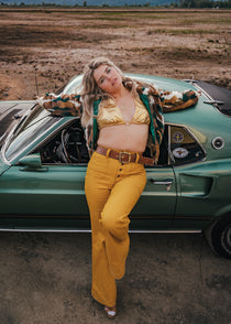 Woman in a gold top and yellow pants standing next to a vintage car in an open field wearing soul sister since 1969.