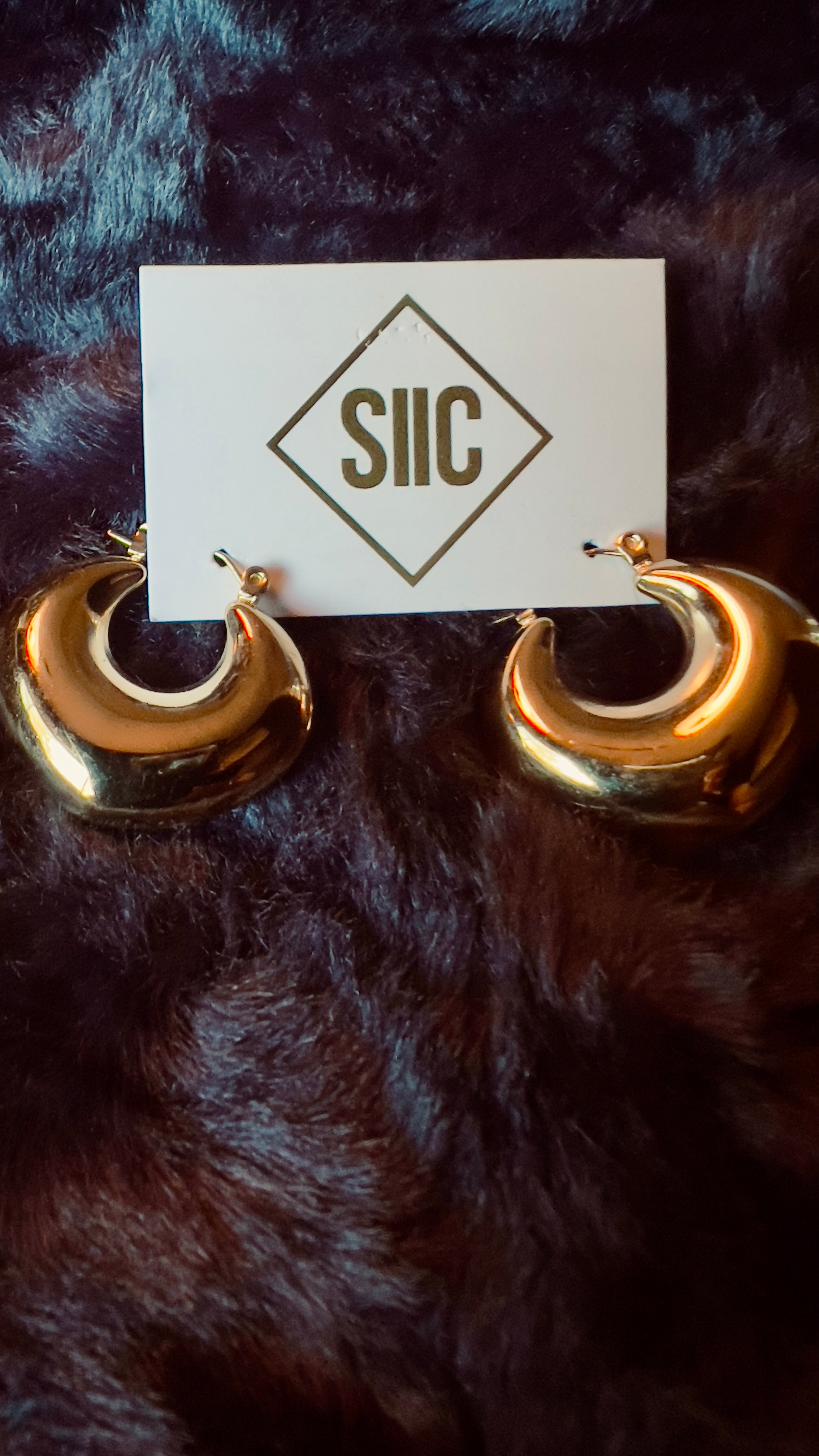 Gold hoop earrings on a dark fur surface with a card displaying 'SIIIC'.