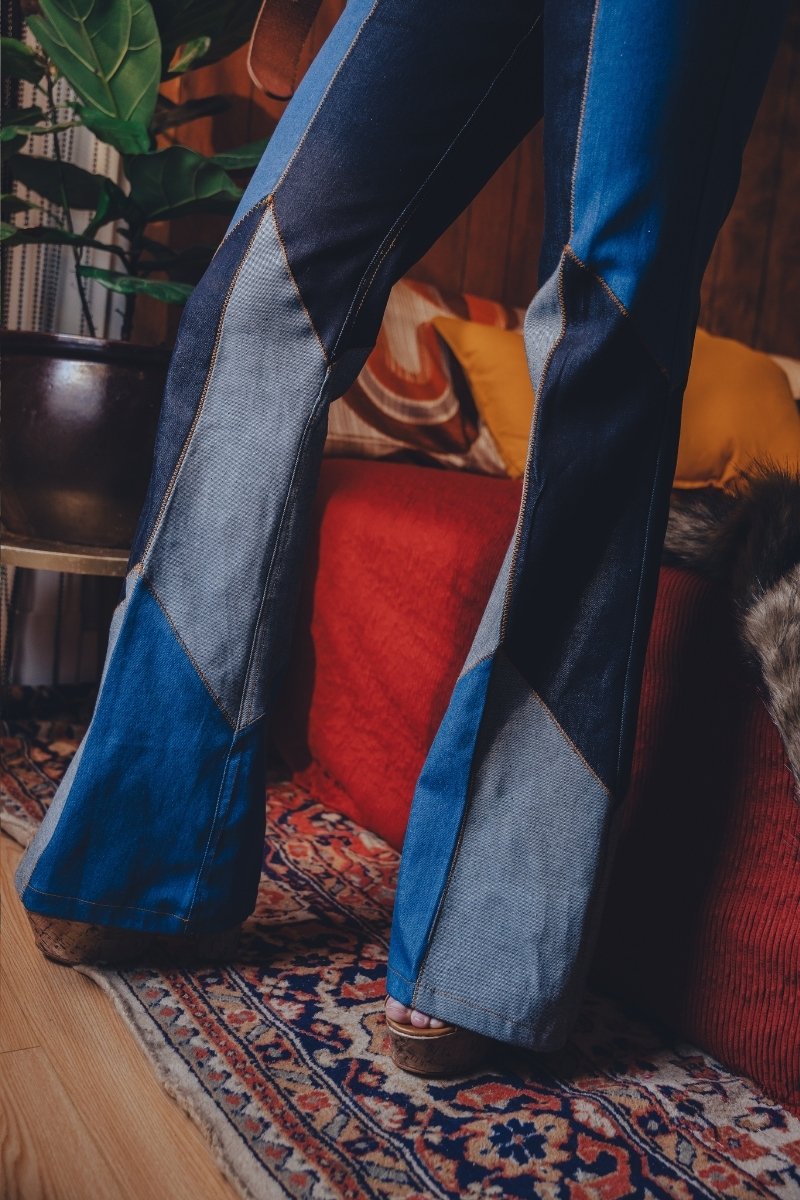 Person wearing blue patchwork pants standing on a patterned rug with a colorful pillow in the background.