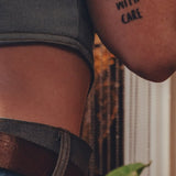 Person wearing a tattoo on their arm with the text 'HANDLE WITH CARE' against a blurred background.