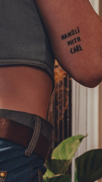 Person wearing a tattoo on their arm with the text 'HANDLE WITH CARE' against a blurred background.