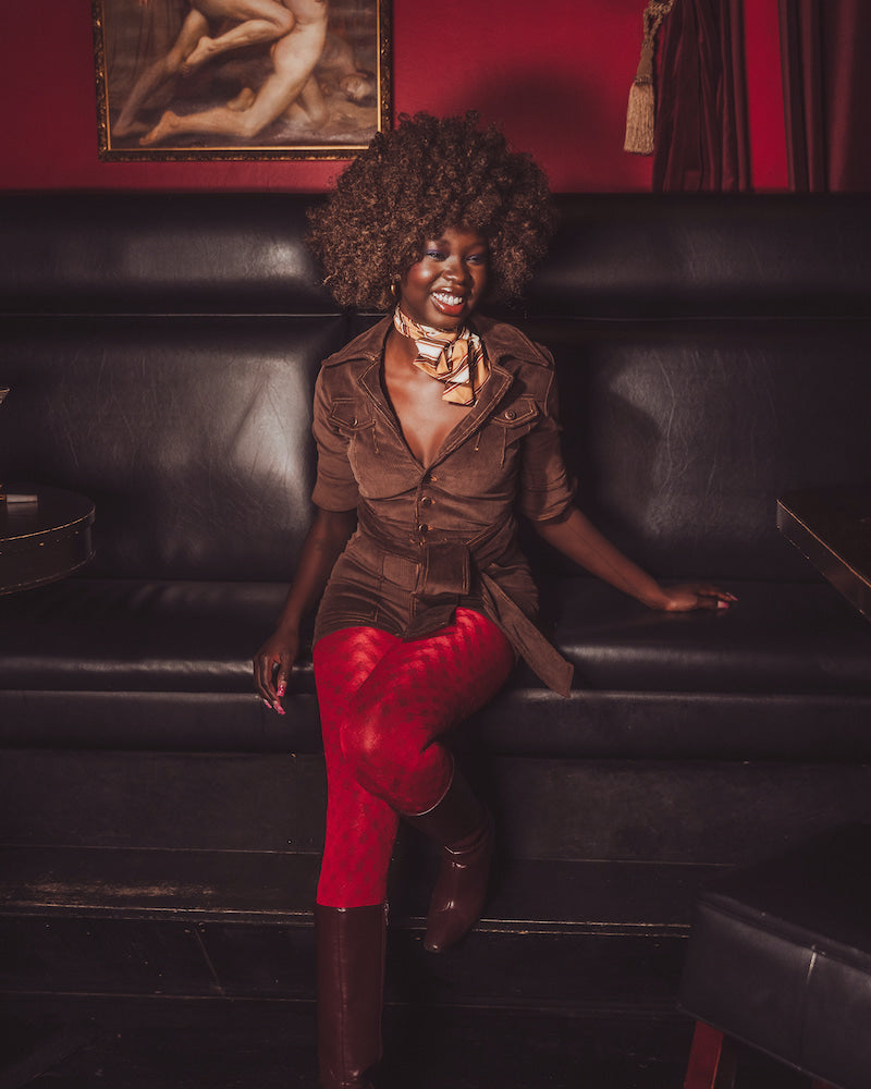 Woman in a brown jacket and red pants sitting on a black leather couch with a red wall and painting in the background.