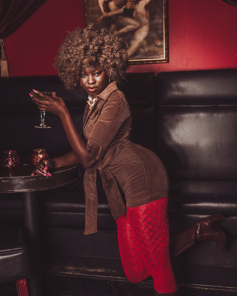 Woman in a brown top and red pants holding a glass in a dimly lit room with a painting on the wall.