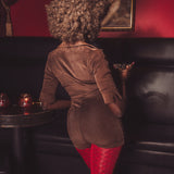 Person in a brown jacket and red pants sitting on a black leather couch with candles and a painting in the background.